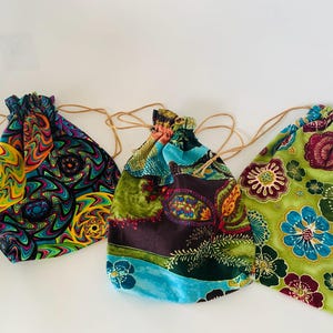 May include: Three drawstring bags with colorful fabric patterns. One bag has a swirling psychedelic design, another features a paisley and floral mix, and the third displays a floral pattern with blue and burgundy accents. Each bag has a natural-colored drawstring.