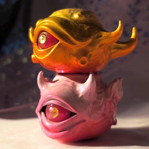 Könnte beinhalten: Zwei gestapelte Skulpturen mit einem fantastischen Design. Die obere Skulptur ist goldfarben, mit strukturierter Oberfläche, Hörnern und einem großen Auge mit gelber Iris. Die untere Skulptur ist rosa, mit ähnlichen Merkmalen. Beide haben ein einzigartiges, künstlerisches Design.