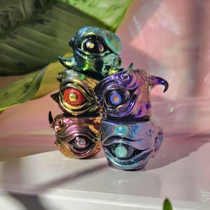 May include: Five small, iridescent, eye-shaped figurines stacked on top of each other. The figurines are made of resin and have a variety of colors, including green, purple, blue, and gold. Each figurine has a different eye design.