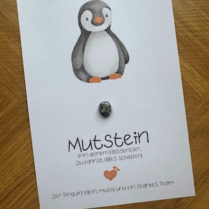 May include: A white card featuring a watercolor illustration of a penguin, a small grey and white speckled stone, and German text that reads Mutstein von deinem Klassentier. Du kannst alles Schaffen! Der Pinguin: Klein, mutig und ein starkes Team.
