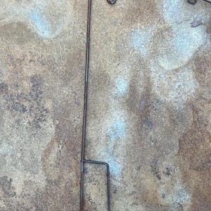 May include: A weathered, dark brown metal shepherd's hook with a curved top for hanging items. The hook is supported by a vertical pole and a two-pronged base for ground insertion. Ideal for outdoor decor.