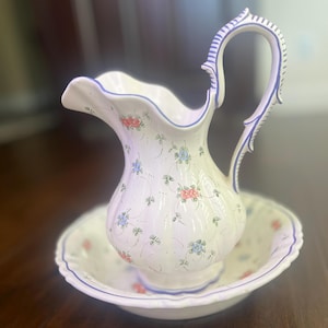 Puede incluir: Un juego de jarra y palangana de cerámica blanca. La jarra tiene un asa y un pico curvos, con un borde festoneado. Ambas piezas están decoradas con pequeños motivos florales en rosa y azul, y delineadas con ribetes azules. El conjunto está sobre una superficie de madera oscura.