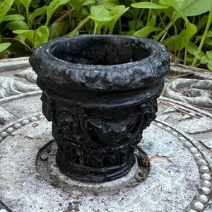 May include: A black, ornate planter with a textured surface. The planter has a wide, rounded rim and a detailed, sculpted design around the base. The planter is sitting on a weathered, gray surface.