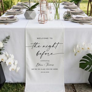 May include: An outdoor wedding reception table set for a celebration. The table is covered with a white tablecloth and decorated with floral arrangements, candles, and place settings. A banner reads "Welcome to the night before".