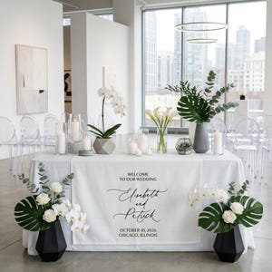 Peut inclure: Une table de réception de mariage est décorée de fleurs blanches, de bougies et de verdure. La table est recouverte d'une nappe blanche et porte une bannière avec l'inscription "Bienvenue à notre mariage", les noms Elisabeth et Patrick, et la date du 10 octobre 2026, à Chicago, Illinois.