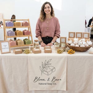 May include: A display of natural soaps and bath products at a craft fair. Soaps in various colors are arranged on a wooden rack. The table features a sign that reads "Vegan & Cruelty-Free" and a banner with the "Bloom & Bare Natural Soap Co." logo.