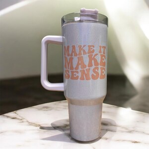 May include: A pearlescent tumbler with a white handle and a clear lid. The tumbler has the words "MAKE IT MAKE SENSE" printed in orange. The tumbler is sitting on a marble surface.