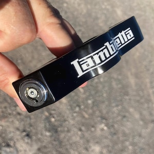 May include: A black Lambretta scooter lock with white lettering. The lock features a keyhole and the Lambretta logo. The lock is held in a hand.