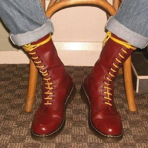 Może przedstawiać: Para czerwonych skórzanych butów z żółtymi sznurowadłami. Buty nosi osoba w niebieskich dżinsach.