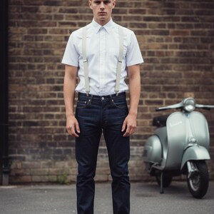 May include: A man wearing a white short-sleeve button-down shirt with light gray suspenders, dark blue jeans, and red shoes. A silver scooter is parked in the background against a brick wall.