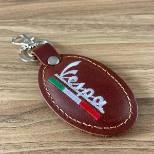 以下が含まれることがあります： 白で刺繍された「Vespa」の文字が入った茶色の革製キーホルダー。 イタリアの国旗の色とシルバーの留め金が付いています。 楕円形の革片は、明るい茶色の糸でステッチされています。