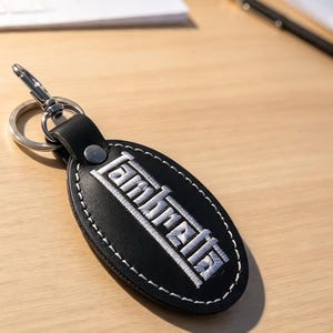 Può includere: Un portachiavi ovale in pelle nera con la scritta ricamata in bianco Lambretta. Il portachiavi presenta cuciture bianche a contrasto lungo i bordi, un rivetto in metallo argentato e un anello circolare con chiusura a moschettone.