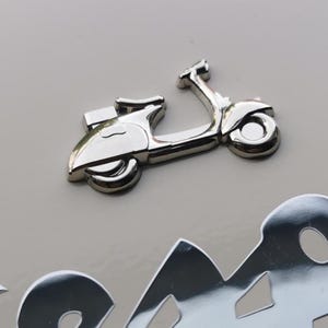 以下が含まれることがあります： 光沢のあるシルバーカラーのスクーターエンブレム。スクーターは、ハンドルバー、シート、ホイールなど、詳細なデザインが施されています。エンブレムは明るいグレーの背景に配置され、スクーターの形状を際立たせています。