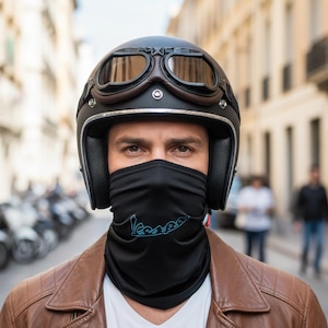 May include: A black motorcycle helmet with brown goggles and a black face covering featuring the Vespa logo in teal. The person is wearing a brown leather jacket and a white shirt.