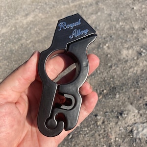 Puede incluir: Una multiherramienta de metal gris oscuro con el texto "Royal Alloy" en plata. La herramienta tiene un abrebotellas, un anillo y un diseño curvo.