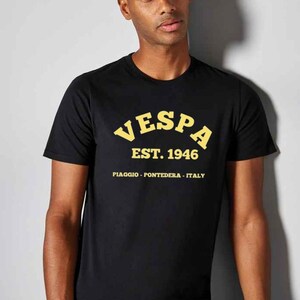 May include: Black crew neck t-shirt with the word "VESPA" in yellow arched lettering, along with "EST. 1946" and "PIAGGIO - PONTEDERA - ITALY" in yellow. The shirt is worn by a person.