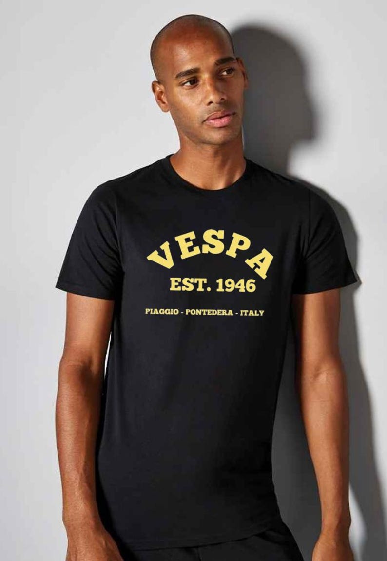 May include: Black crew neck t-shirt with the word "VESPA" in yellow arched lettering, along with "EST. 1946" and "PIAGGIO - PONTEDERA - ITALY" in yellow. The shirt is worn by a person.
