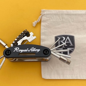 Op de afbeelding: Een zwarte multitool met de tekst "Royal Alloy" in het wit, met diverse opzetstukken. Naast de tool ligt een canvas tas met het "RA" logo. De achtergrond is effen geel.