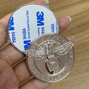 May include: A silver-colored Vespa emblem with a bee design, featuring the text "VESPA" and "PIAGGIO ITALIA". The emblem is round and has a 3M double-sided adhesive backing for easy application.
