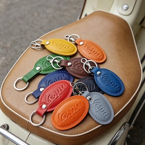 Può includere: Una collezione di portachiavi in pelle di forma ovale in vari colori, tra cui giallo, arancione, verde, marrone, blu, rosso e grigio. Ogni portachiavi presenta il logo Vespa in rilievo, cuciture bianche e un anello in metallo argentato.