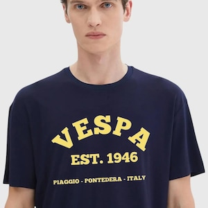 May include: Navy blue t-shirt with the word "VESPA" in yellow arched lettering, along with "EST. 1946" and the location "PIAGGIO - PONTEDERA - ITALY". The shirt is short-sleeved.