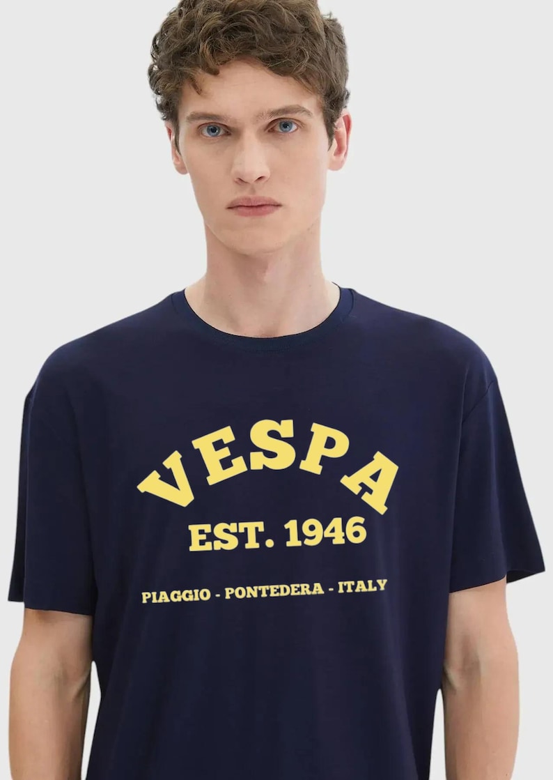 May include: Navy blue t-shirt with the word "VESPA" in yellow arched lettering, along with "EST. 1946" and the location "PIAGGIO - PONTEDERA - ITALY". The shirt is short-sleeved.