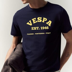 May include: Navy blue t-shirt with the word "VESPA" in yellow arched lettering, along with "EST. 1946" and "PIAGGIO - PONTEDERA - ITALY" printed below. The shirt is short-sleeved.