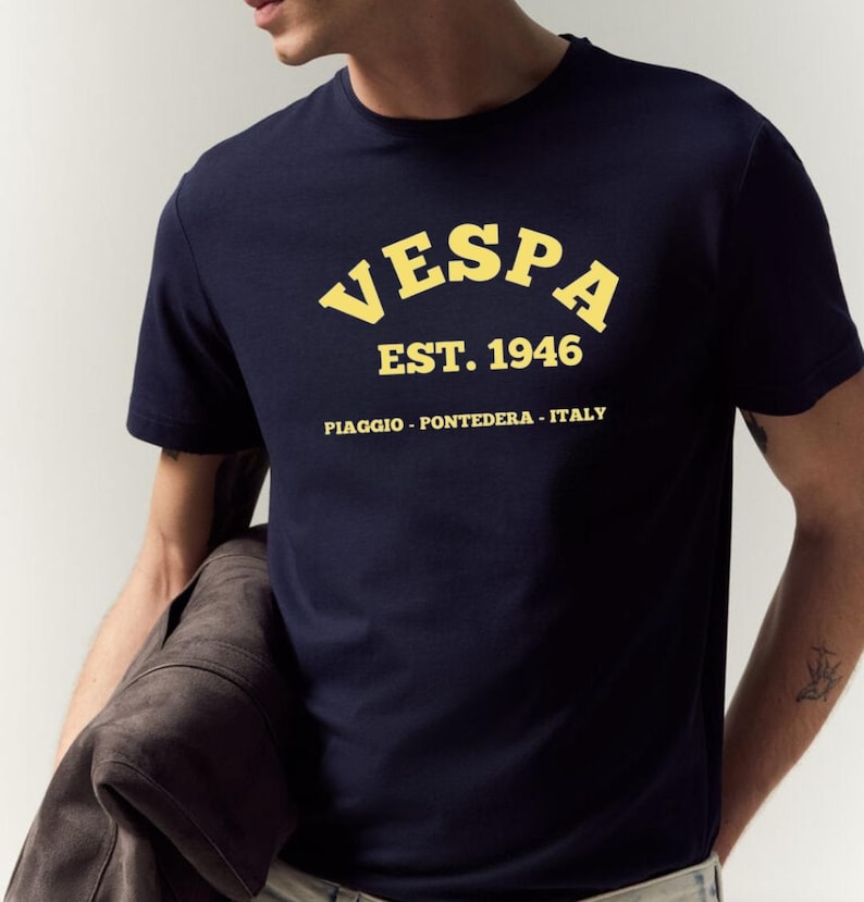May include: Navy blue t-shirt with the word "VESPA" in yellow arched lettering, along with "EST. 1946" and "PIAGGIO - PONTEDERA - ITALY" printed below. The shirt is short-sleeved.