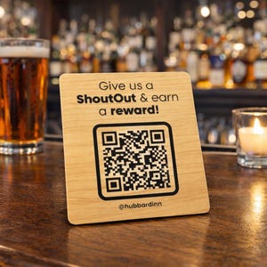 May include: A wooden sign with the text "Give us a ShoutOut & earn a reward!" and a QR code. A glass of beer and a lit candle are also on the bar. The sign is for @hubbardinn.