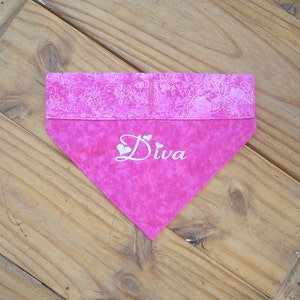 Op de afbeelding: Roze hondenbandana met een witte bloemenrand en het woord "Diva" geborduurd in wit met twee kleine hartjes.