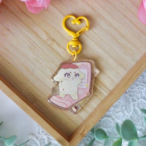 May include: A yellow heart-shaped keychain with a clear charm featuring a cartoon dog wearing a red beret and holding a pink cell phone. The charm is on a wooden surface.