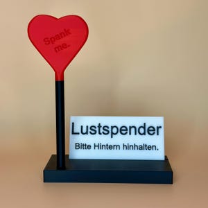 Könnte beinhalten: Ein neuartiger Artikel mit einem roten herzförmigen Paddel mit dem Text "Spank me". Ein schwarz-weißes Schild liest "Lustspender. Bitte Hintern hinhalten."