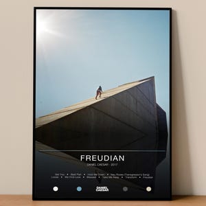 Puede incluir: Impresión artística enmarcada que muestra a una persona subiendo una estructura de hormigón bajo un cielo brillante. La impresión incluye el texto "FREUDIAN" y "DANIEL CAESAR - 2017", junto con una lista de títulos de canciones y el nombre del artista.