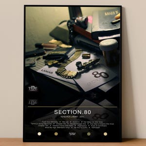 May include: A framed poster featuring the album "Section.80" by Kendrick Lamar. The poster includes the album title, artist name, and a tracklist. Gold jewelry and other items are visible on a table in the background.