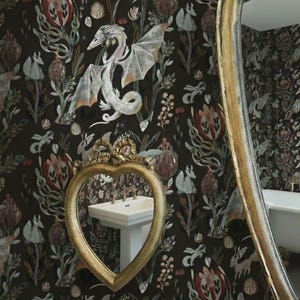 May include: A bathroom scene featuring a heart-shaped mirror with a gold frame, reflecting a white sink. The wallpaper is dark with a floral and fantasy design, including a dragon. A large, oval mirror with a gold frame is also visible.