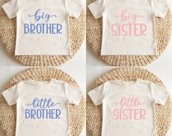 May include: Four white t-shirts with different text printed on them. The text on the shirts reads "big brother", "big sister", "little brother", and "little sister". The text is in a cursive font and is printed in blue and pink.