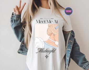 May include: Ivory-colored t-shirt featuring a Cinderella-themed graphic. The design includes the text "Meet Me At Midnight" with an illustration of Cinderella's profile. The shirt has a round neck and short sleeves.
