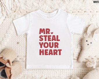 May include: White short-sleeved t-shirt with the text "MR. STEAL YOUR HEART" in red. The shirt is displayed on a cream-colored surface with a stuffed bunny and baby shoes.