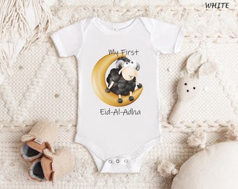 May include: White baby onesie with a cartoon sheep on a crescent moon. The sheep is black and white with a tan face. Text on the onesie reads "My First Eid-Al-Adha".