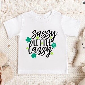 May include: White short-sleeved t-shirt with the text "sassy little lassy" in black script, accented with green shamrocks. The shirt is designed for children and is displayed on a textured surface.