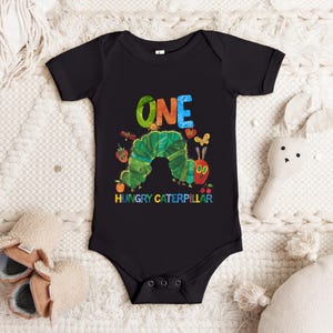 May include: Black baby bodysuit with a colorful graphic of a green caterpillar, the word "ONE" in rainbow colors, and the text "HUNGRY CATERPILLAR". The bodysuit is short-sleeved and has snap closures.