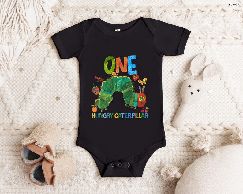 May include: Black baby bodysuit with a colorful graphic of a green caterpillar, the word "ONE" in rainbow colors, and the text "HUNGRY CATERPILLAR". The bodysuit is short-sleeved and has snap closures.