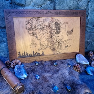 Op de afbeelding: Een houten ingelijste kaart van Midden-aarde uit The Lord of the Rings. De kaart is gegraveerd met de namen van verschillende locaties, waaronder De Shire, Mordor en Gondor. De lijst is donkerbruin met een decoratieve rand.