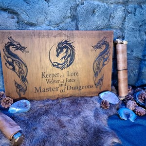 Op de afbeelding: Een houten bord met een drakenontwerp en de tekst "Keeper of Lore, Weaver of Fates and Master of Dungeons".