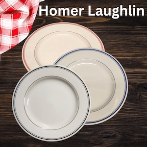 May include: Three white ceramic plates with blue and red trim. The plates are stacked on top of each other. The plates are on a wooden surface. The text "Homer Laughlin" is visible in the image.
