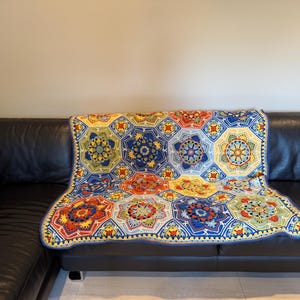 Pode incluir: Uma manta de crochê colorida e feita à mão com um padrão floral geométrico em tons de azul, amarelo, vermelho e verde. A manta está sobre um sofá de couro preto, mostrando seu design intrincado e artesanato.