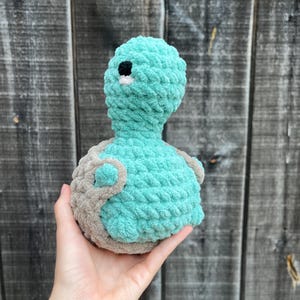 May include: A handmade crochet turtle plushie in shades of teal and gray. The turtle's head and body are teal, with a black eye and white accent. The shell and flippers are gray. The plushie is soft and textured.