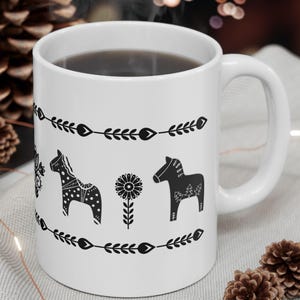 May include: White ceramic mug with a black and white design of stylized horses and flowers. The mug is filled with coffee and has a black handle.