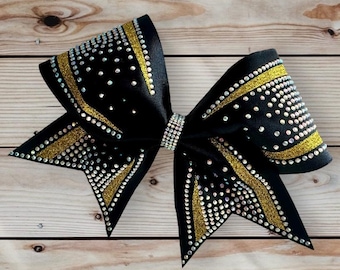 May include: A black cheer bow with gold glitter and rhinestones. The bow is tied in a large, symmetrical shape.