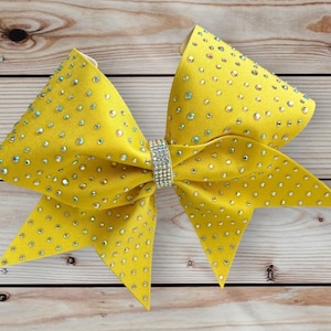 May include: A large yellow cheer bow with a rhinestone design. The bow is made of satin fabric and has a center piece with clear rhinestones.